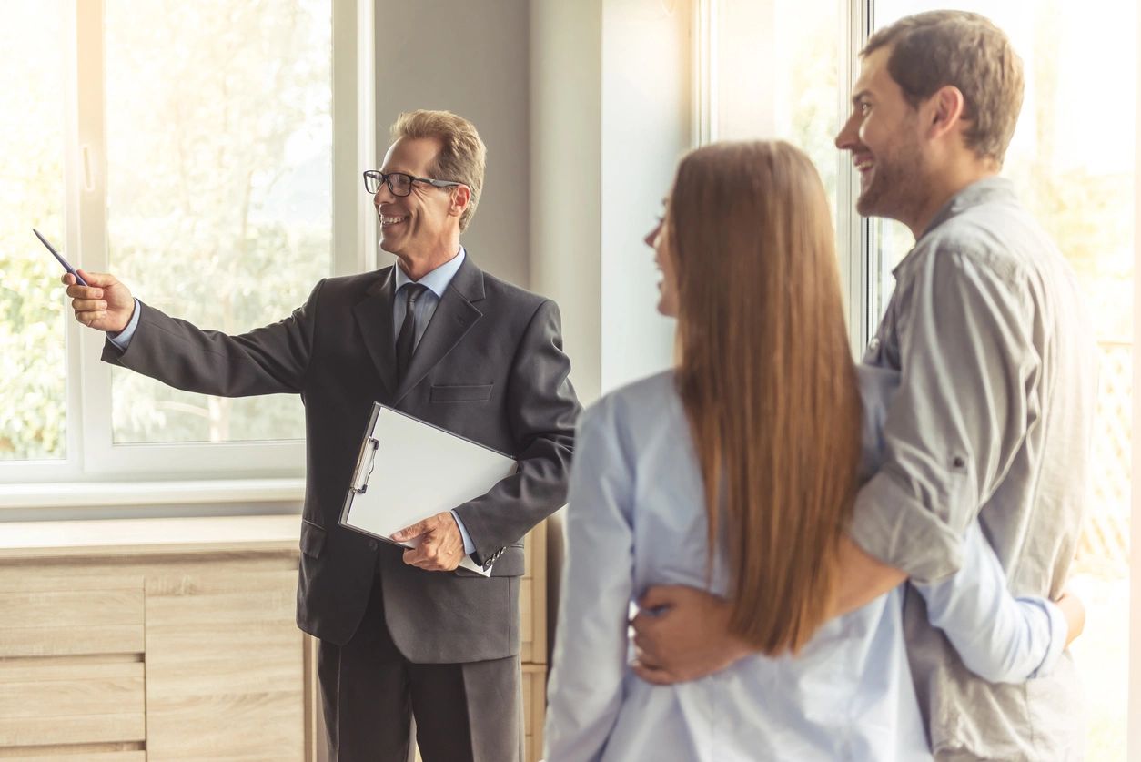 Handsome middle aged realtor in classic suit and eyeglasses is holding a notepad and smiling while showing new apartment to young couple (Handsome middle aged realtor in classic suit and eyeglasses is holding a notepad and smiling while showing new ap